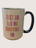 Beige mug with colorful text 'JUST AN OLD HO CARRYING ON' on a light gray background