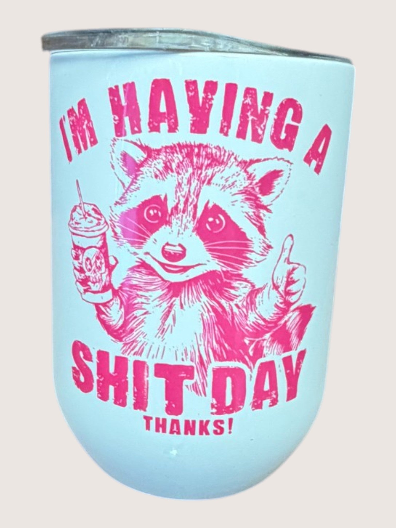 Light blue travel cup/tumbler with pink raccoon graphic and text on a light pink background