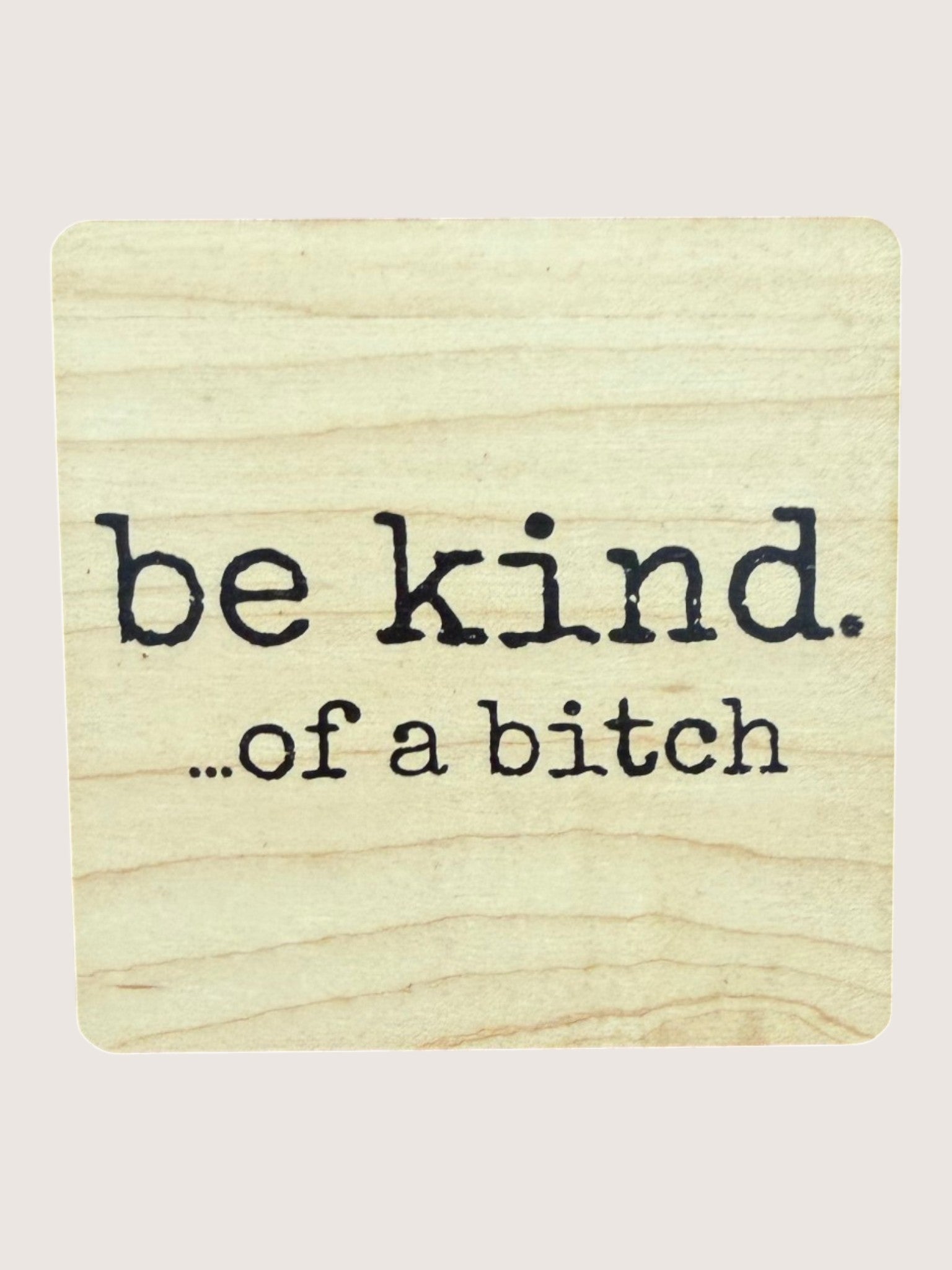 Wooden coaster with 'be kind...of a bitch' text on a light background