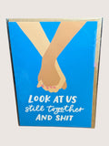 Card with illustration of two hands holding each other and text 'Look at us still together AND SHIT' on a blue background.