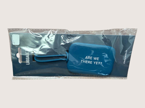 Blue luggage tag with text 'Are we there yet?' in packaging on a light gray background