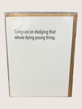 Card with humorous text 'Congrats on dodging that whole dying young thing.' on a white background.