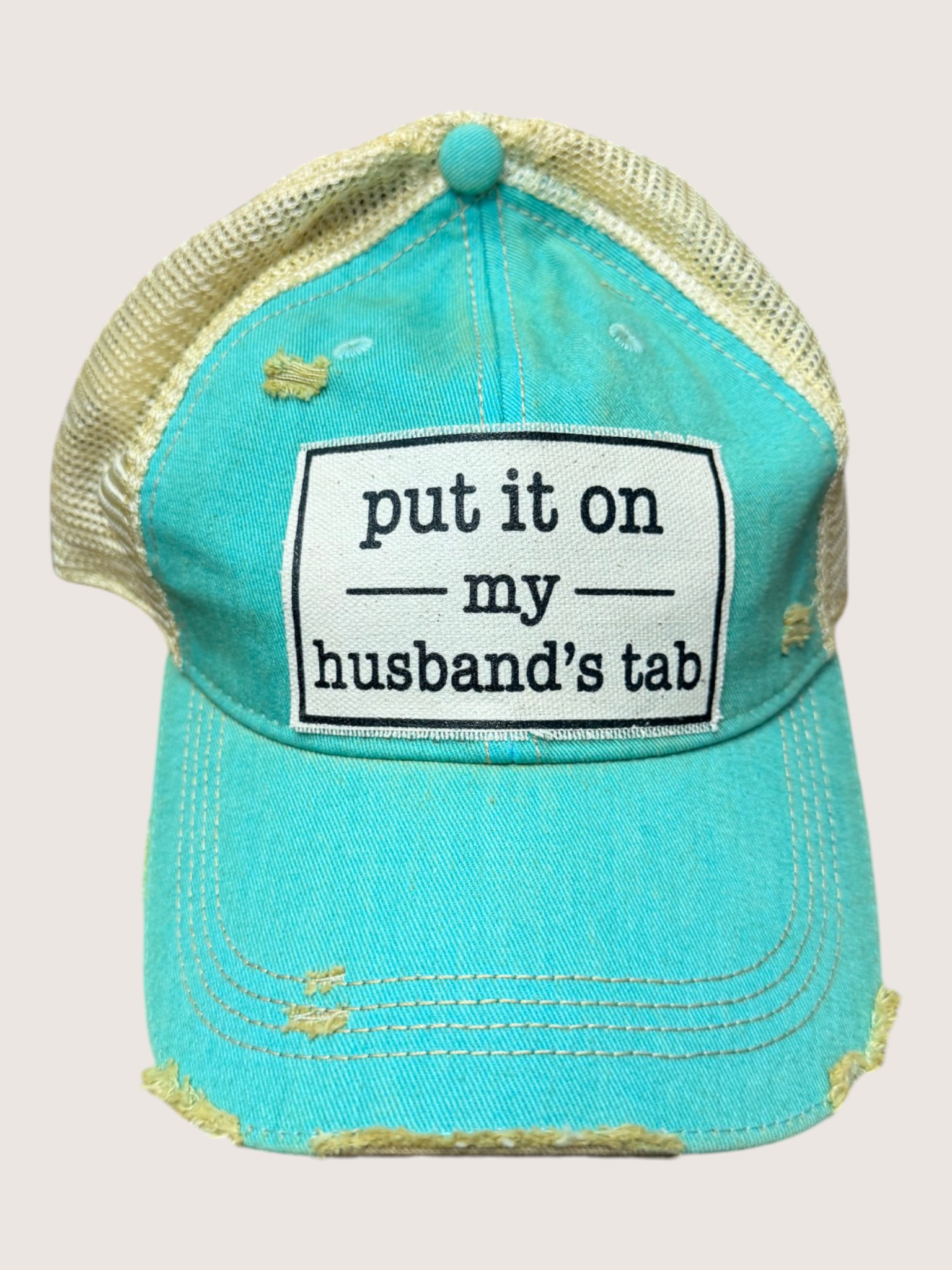 Turquoise cap with text 'put it on my husband's tab' on a light gray background