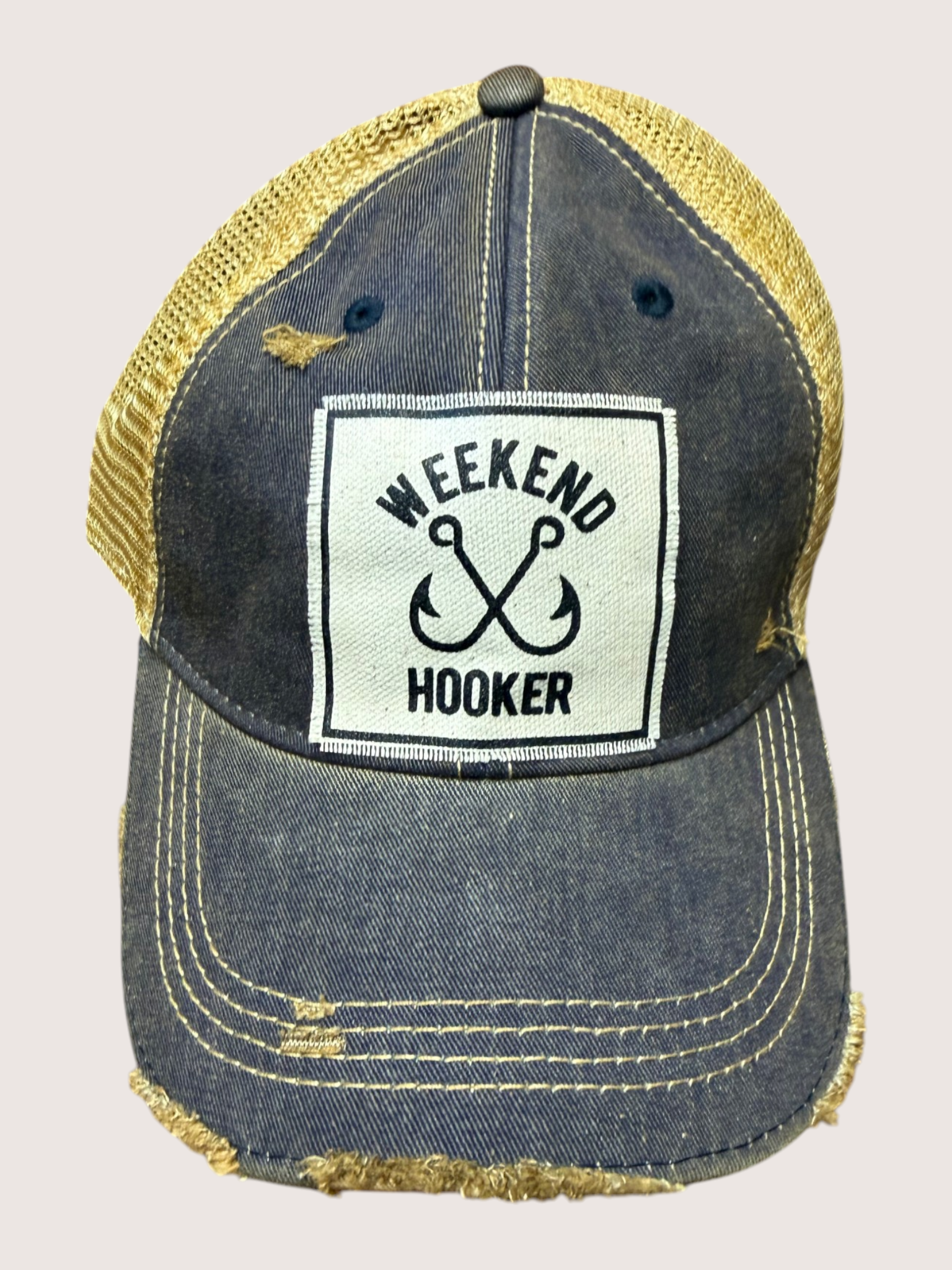 Blue trucker cap with 'Weekend Hooker' patch on a beige background