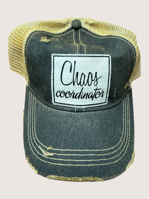 Distressed denim cap with 'Chaos coordinator' patch on a light pink background