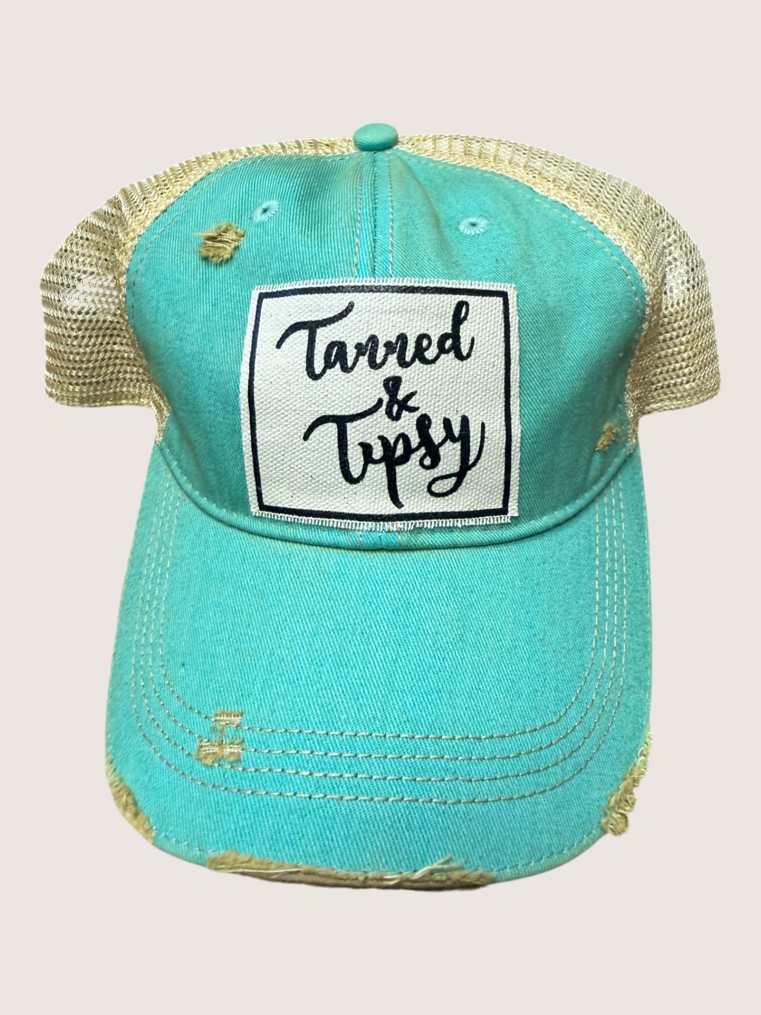 Turquoise cap with 'Tanned & Tipsy' text on a beige background