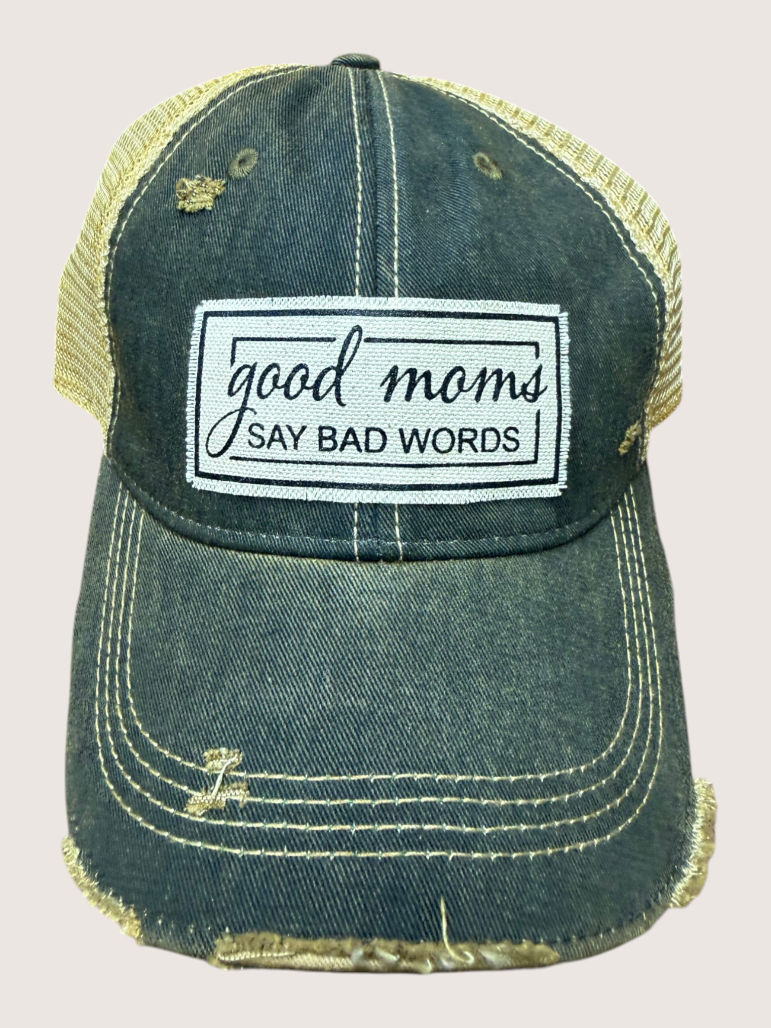 Blue denim cap with yellow mesh back and 'good moms say bad words' patch on a beige background