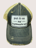 Distressed green trucker hat/cap with a white patch reading 'put it on my boyfriend's tab' on a light pink background.