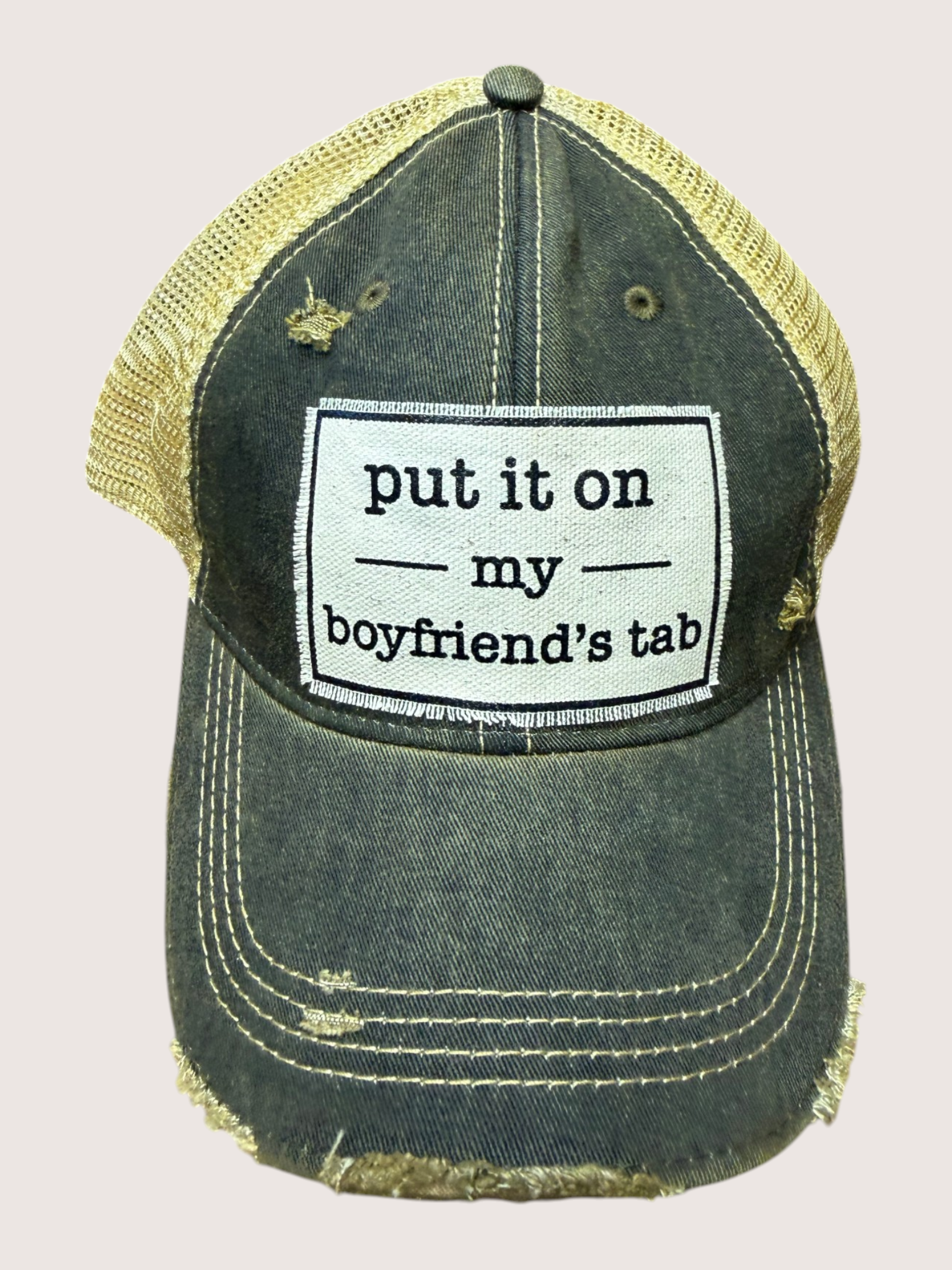 Distressed green trucker hat/cap with a white patch reading 'put it on my boyfriend's tab' on a light pink background.