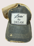 Distressed denim cap with 'Livin' the Dream' patch on a light pink background