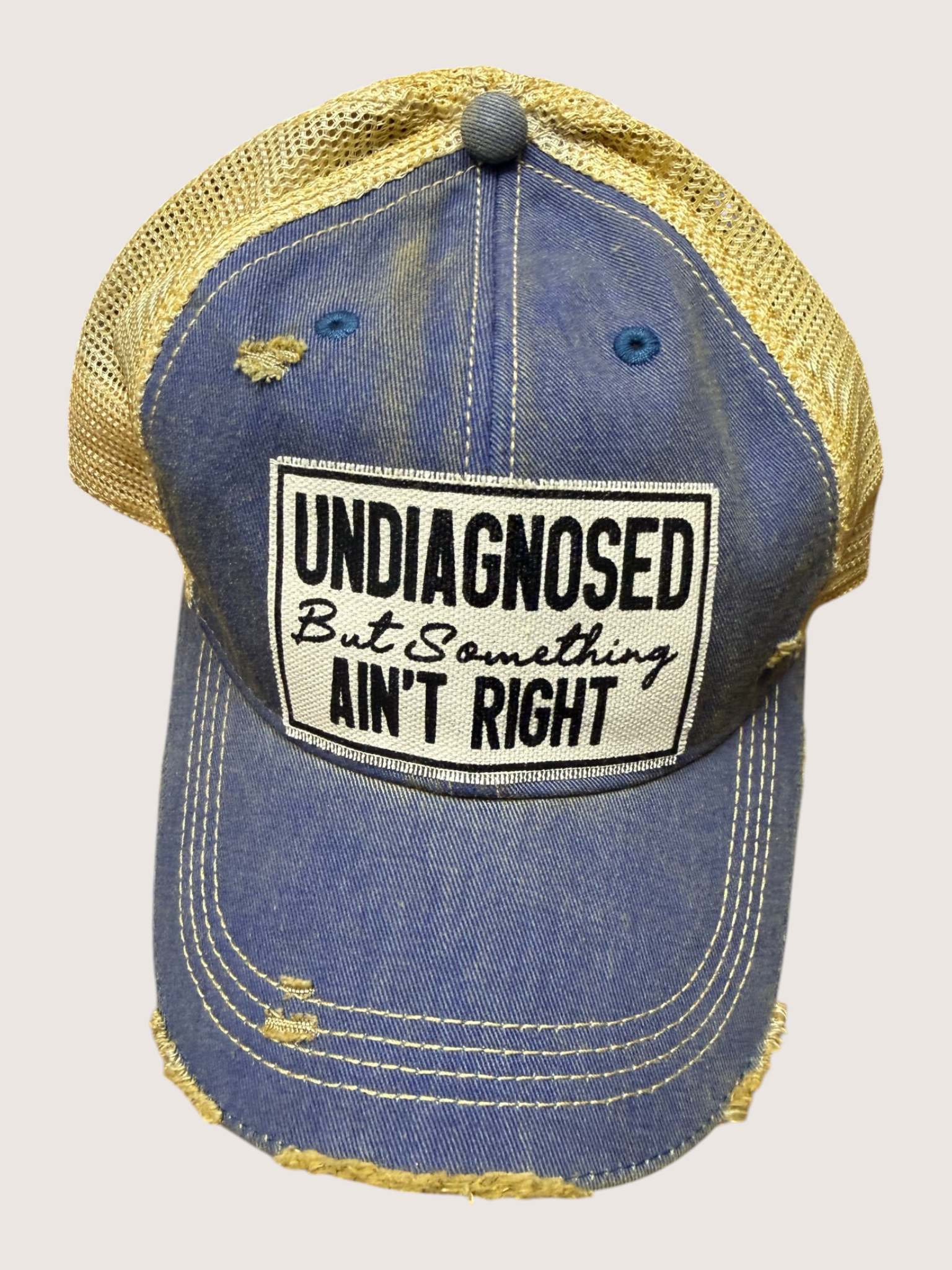 Blue and yellow cap with a patch reading 'undiagnosed But Something Ain't Right' on a beige background