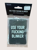 Air freshener with text 'Use Your Fucking Blinker' in packaging on a light gray background