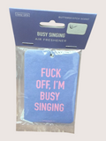 Air freshener with text 'F*ck off, I'm busy singing' in pink on a blue background, packaged in clear plastic with a blue label.