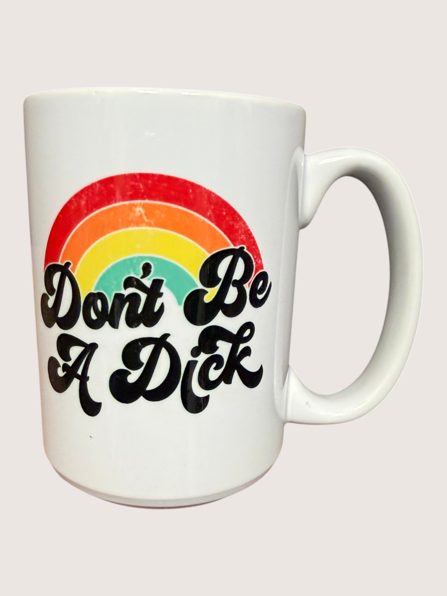 White mug with rainbow and text 'Don't Be A Dick' on a light pink background